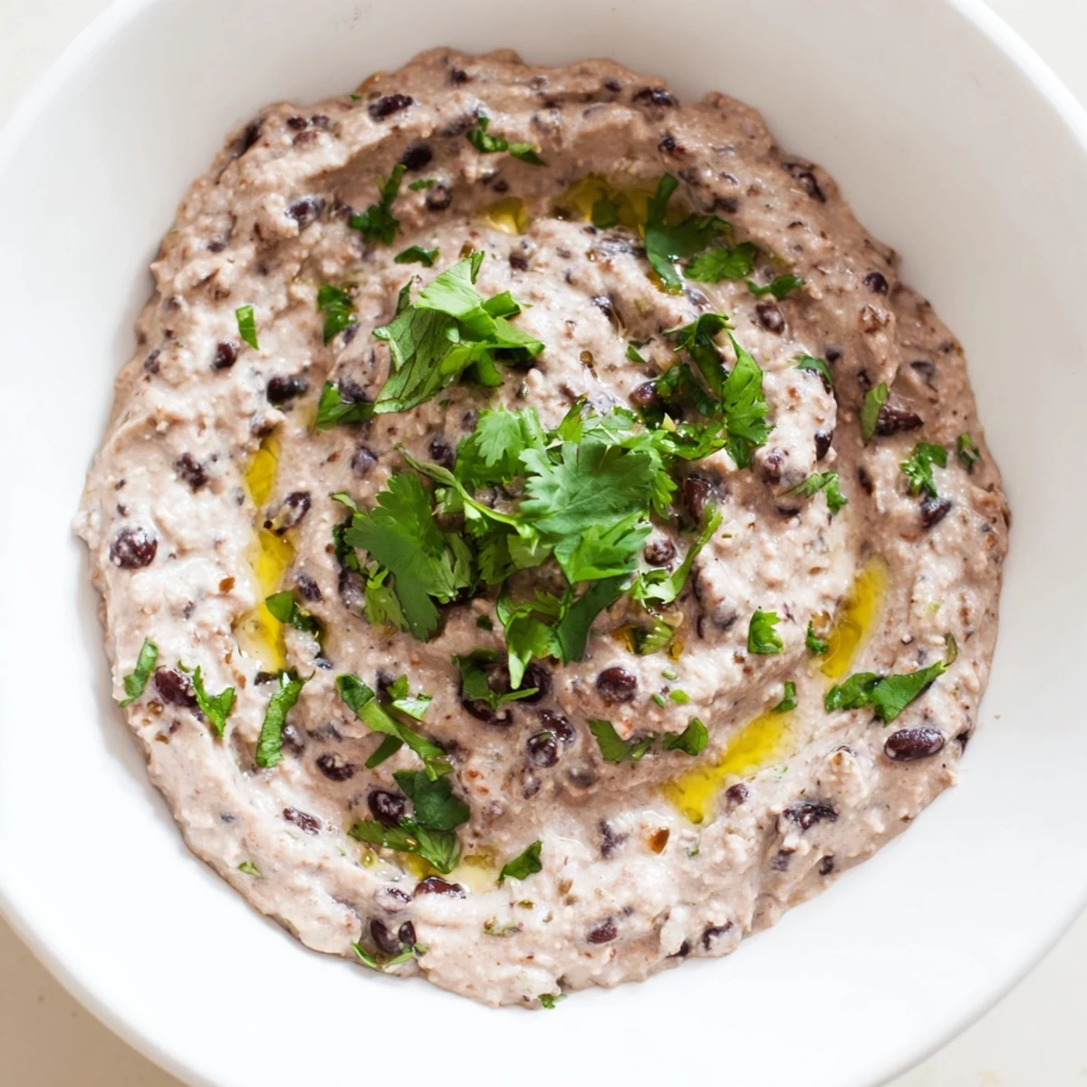 A smooth bowl of spicy black bean dip topped with fresh cilantro, lime wedges, and sliced jalapeños.  