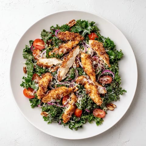 Golden, crispy chicken strips top a hearty kale salad, garnished with cherry tomatoes and toasted pecans for a satisfying, crunchy bite.